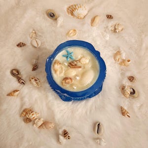 Peut inclure: Un bougeoir bleu rempli de cire crémeuse, décoré de coquillages, de perles et d'une étoile de mer bleue. Divers coquillages sont dispersés autour du bougeoir sur une surface blanche et duveteuse.