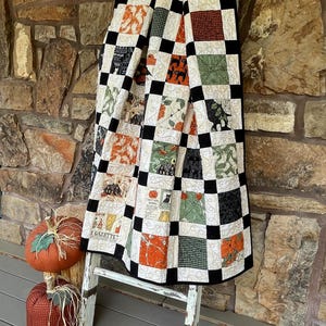 May include: A patchwork quilt featuring a grid of black squares alternating with various Halloween-themed fabric squares in orange, green, and cream. The quilt displays printed motifs of pumpkins, black cats, and text including GAZETTE.