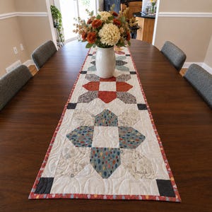 May include: A rectangular quilted table runner featuring a patchwork design with star-shaped motifs in shades of rust orange, blue, and cream. The fabric displays intricate swirling machine quilting patterns and a decorative border with a colorful print.