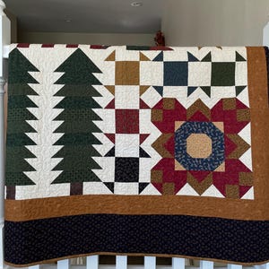 May include: A patchwork quilt featuring a repeating pattern of dark green pine trees, geometric blocks in shades of burgundy, navy blue, mustard yellow, and cream, finished with a wide tan border and a dark navy blue bottom edge.