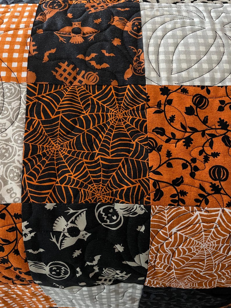 以下が含まれることがあります： ハロウィーンをテーマにした様々な布の正方形を組み合わせたパッチワークキルト。オレンジと黒のクモの巣、白いフクロウとカボチャが描かれた黒い布、黒いツル模様のオレンジ色の布、灰色のカボチャの輪郭が描かれた白い布などのパターンが含まれています。