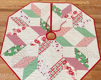 May include: An octagonal quilted Christmas tree skirt featuring a patchwork design of white, red, and green fabrics. The pattern includes prints of Santa Claus, poinsettias, and plaid motifs, finished with a red fabric border and two red ties.