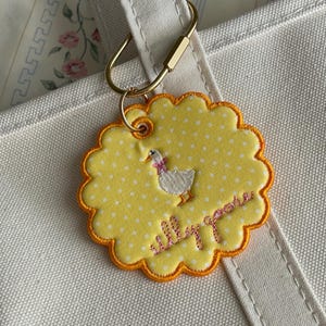 May include: A round, scalloped-edge yellow fabric keychain with white polka dots, featuring an embroidered white goose with a pink bow and the cursive text "silly goose" in pink thread, attached to a gold-toned metal ring on a white canvas bag.