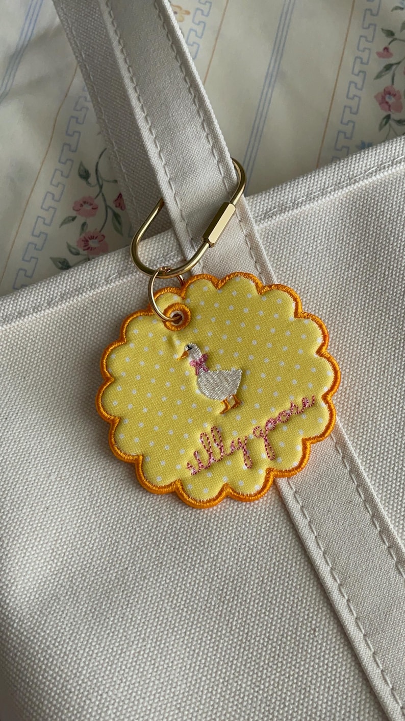 May include: A round, scalloped-edge yellow fabric keychain with white polka dots, featuring an embroidered white goose with a pink bow and the cursive text "silly goose" in pink thread, attached to a gold-toned metal ring on a white canvas bag.