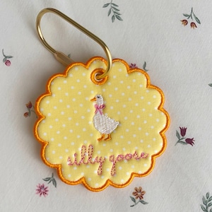 May include: A round, scalloped-edge yellow keychain with white polka dots, featuring an embroidered white goose with a pink bow and the text "silly goose" in pink thread. The keychain is attached to a gold-toned metal ring.