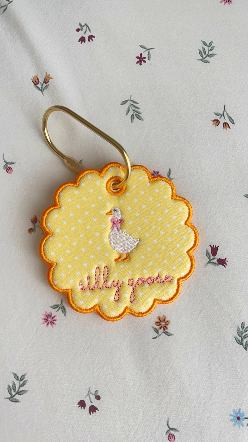 May include: A round, scalloped-edge yellow keychain with white polka dots, featuring an embroidered white goose with a pink bow and the text "silly goose" in pink thread. The keychain is attached to a gold-toned metal ring.
