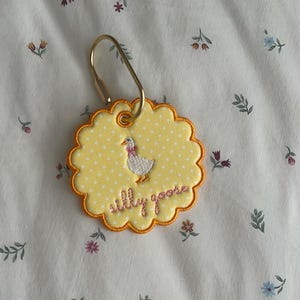 May include: A round, scalloped-edge yellow fabric tag with white polka dots, featuring an embroidered white goose with a pink bow and the text "silly goose" in pink thread. The tag is attached to a gold-toned metal lobster clasp ring.