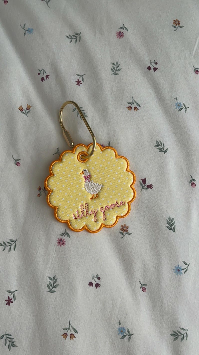 May include: A round, scalloped-edge yellow fabric tag with white polka dots, featuring an embroidered white goose with a pink bow and the text "silly goose" in pink thread. The tag is attached to a gold-toned metal lobster clasp ring.