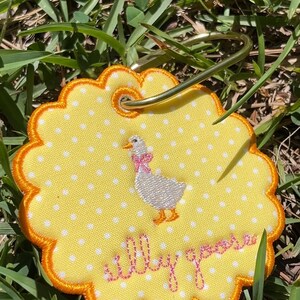 May include: A round, scalloped-edge yellow fabric tag with white polka dots, featuring an embroidered white goose with a pink bow and the text "silly goose" in pink script. A gold-toned metal keyring is attached through a reinforced eyelet.