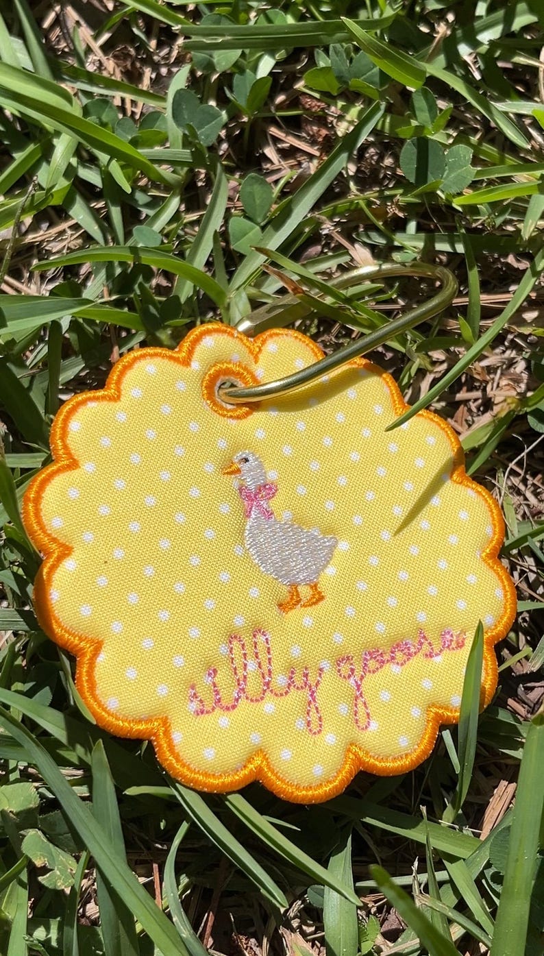 May include: A round, scalloped-edge yellow fabric tag with white polka dots, featuring an embroidered white goose with a pink bow and the text "silly goose" in pink script. A gold-toned metal keyring is attached through a reinforced eyelet.