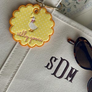 May include: A cream-colored canvas tote bag featuring the embroidered monogram SDM in dark brown thread. Attached to the handle is a round, scalloped yellow patch with white polka dots, a white goose illustration, and the text silly goose.