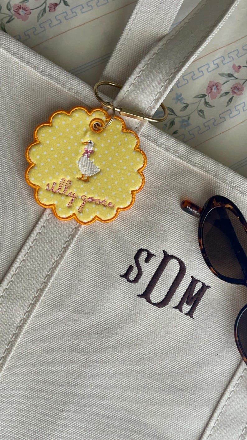May include: A cream-colored canvas tote bag featuring the embroidered monogram SDM in dark brown thread. Attached to the handle is a round, scalloped yellow patch with white polka dots, a white goose illustration, and the text silly goose.