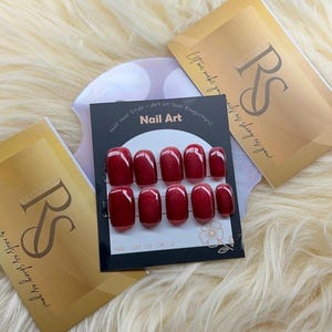 Puede incluir: Un conjunto de uñas postizas rojas con acabado brillante, presentado en una tarjeta negra con el texto "Nail Art". La tarjeta está rodeada de tarjetas doradas con el logotipo "RS" y la frase "Let me make your nails as shiny as you!"