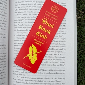 May include: A red bookmark with gold lettering and a yellow hibiscus flower graphic rests on an open book. The bookmark reads "Diwi Book Club" and includes text in Hawaiian. The book's title is "The Killing Spell."