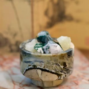 May include: A small, cylindrical stone bowl with a marbled pattern in shades of green, cream, and dark brown. The bowl is filled with several raw, translucent, and opaque mineral stones in white and deep green hues. The text WU LI Marble is visible.