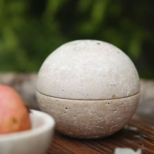 May include: A spherical beige stone container with a porous, textured surface and a horizontal seam dividing the top and bottom halves. The top of the image features the text WU LI Marble with small pink and gold vase icons above the lettering.
