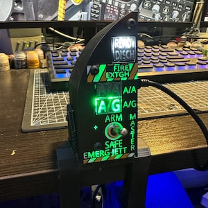 Puede incluir: Un dispositivo electrónico negro con texto verde y blanco, incluyendo "READY DISCH" y "FIRE EXTGH". El dispositivo tiene un interruptor basculante y está montado en un soporte negro. Un cable está conectado al dispositivo.