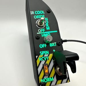 May include: A black control panel featuring illuminated green text labels including IR COOL, ORIDE, NORM, OFF, HMD, BRT, SPIN, RCVY, and NORM. The device includes a metal toggle switch, a rotary knob, a guarded switch, and yellow and black hazard stripes.