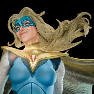 Op de afbeelding: Een gedetailleerd superheldenbeeldje met lang, golvend blond haar, een blauw masker, een gouden kraag met een rood-wit embleem en een blauw-wit gestructureerd pak. Het personage heeft een lichte glimlach en kijkt omhoog.