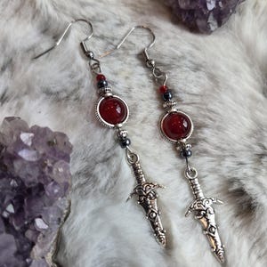May include: A pair of dangle earrings featuring silver-toned sword charms, round deep red beads set in ornate silver frames, and small dark metallic accent beads. The earrings hang from silver-toned hooks and include decorative metal spacers.
