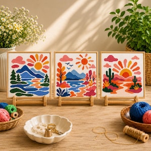 May include: Three square cross-stitch embroideries in wooden frames, each depicting a colorful landscape with a sun, clouds, and terrain. The left shows mountains and water, the center features trees and hills, and the right displays cacti and desert.