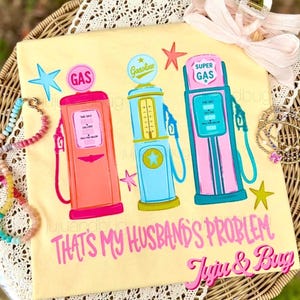 Pode incluir: Camiseta amarelo claro com três bombas de gasolina retrô em rosa, azul e turquesa, com estrelas. O texto diz "THAT'S MY HUSBAND'S PROBLEM" e "Juju & Bug".