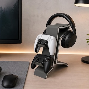 May include: A gray and white gaming headset and controller stand. The stand holds a black headset, two game controllers, and is on a wooden desk. A computer monitor, keyboard, and mouse are also visible.