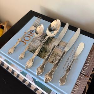 Könnte beinhalten: Ein Set aus sechs silberfarbenen Metallbestecken, bestehend aus einer Gabel, einem kleinen Löffel, einem Schlitzlöffel, zwei Messern und einem Buttermesser, alle mit verzierten Blumenreliefgriffen, angeordnet auf einem hellblauen Buch mit dem Titel Joie.
