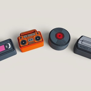 Pode incluir: Um conjunto de quatro teclas retangulares e circulares com designs de eletrônicos retrô. A coleção inclui uma fita VHS cinza-escura, um boombox laranja brilhante, um disco de vinil cinza-escuro com centro vermelho e um console de videogame.