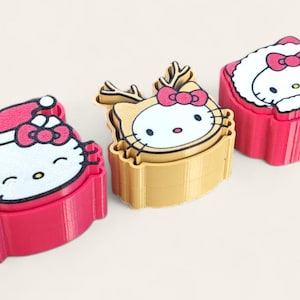 May include: Three plastic cookie cutters featuring Hello Kitty designs. The left cutter is pink with a Santa hat, the center is gold with reindeer antlers, and the right is pink with a winter hood. Each cutter has a detailed face graphic.