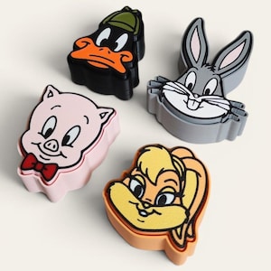 Peut inclure: Un ensemble de quatre emporte-pièces en forme de personnages : Daffy Duck en noir, Bugs Bunny en gris, Porky Pig en rose clair avec un nœud papillon rouge, et Lola Bunny en orange clair. Chaque pièce présente un contour et des détails faciaux.