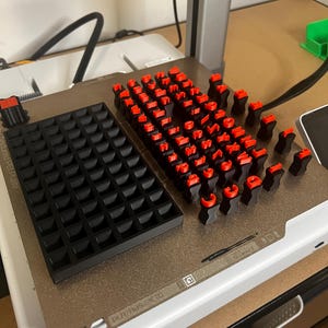 Puede incluir: Una placa de construcción de impresora 3D con una bandeja de rejilla negra y una colección de caracteres individuales de letras y números en rojo y negro. La placa metálica texturizada incluye etiquetas de texto y marcas.