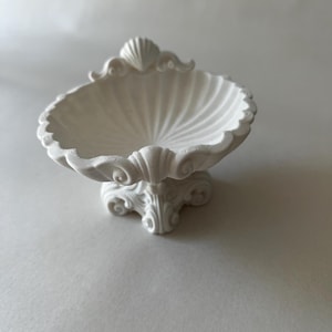May include: A matte white shell-shaped bowl with a scalloped edge and a raised seashell motif at the top. The bowl is supported by a decorative pedestal base featuring intricate scrollwork details, creating a cohesive and textured appearance.