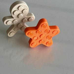 May include: A beige gingerbread person-shaped figure and an orange star-shaped figure, both featuring a textured surface with raised circular studs across their front faces. These two distinct shapes are positioned upright on a smooth surface.