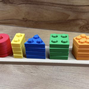 May include: A wooden tray holds five colorful, stacked geometric shapes: a red cylinder, yellow rectangle, blue triangle, green square, and orange hexagon. Each shape has raised circular studs on top, resembling building blocks.