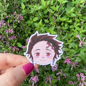 Könnte beinhalten: Ein gestanzter Vinyl-Aufkleber mit einem stilisierten Anime-Charakterkopf mit dunkelbraunem Haar, einer roten Stirnmarkierung, großen roten Augen und kleinen rechteckigen Ohrringen mit einem sonnenartigen Design.