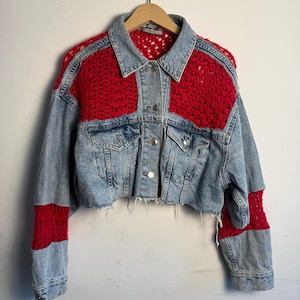 May include: Cropped denim jacket with red crochet detailing on the shoulders and sleeves. The jacket has a classic collar, button-up front, and raw hem. The denim is a light wash blue.
