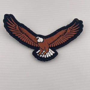 May include: An embroidered patch featuring a bald eagle in flight with wings spread wide. The eagle has a brown body, white head and tail feathers, and a yellow beak. The entire design is outlined with a thick black border on a fabric base.