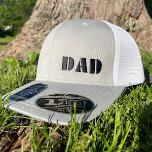 Peut inclure: Casquette de camionneur grise et blanche avec le mot "DAD" brodé en noir. La casquette a une visière incurvée et un dos en filet. La casquette est posée dans l'herbe verte.