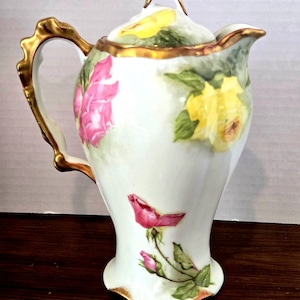 May include: White ceramic pitcher with a lid, adorned with pink and yellow rose floral designs. The handle and rim are accented with gold trim. The pitcher has a bulbous base and a slender neck.