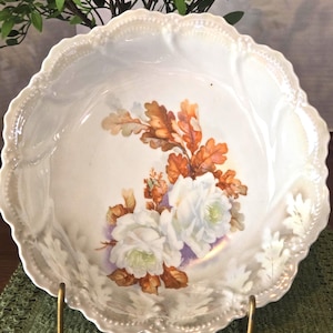 May include: A round porcelain bowl with an iridescent finish and a scalloped, beaded edge. The center features a painted design of two white roses surrounded by clusters of brown and orange oak leaves, set against a soft purple background.