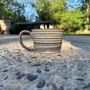 May include: A ceramic mug featuring a horizontal ribbed texture with a variegated glaze in shades of sage green, deep blue, and earthy brown. The mug has a wide, rounded handle and a slightly flared rim, showcasing a handcrafted appearance.