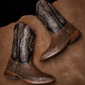 Op de afbeelding: Een paar bruine cowboylaarzen met een slangenpatroon op het onderste deel en een donkerbruin leren bovenwerk. De laarzen hebben decoratieve stiksels en een vierkante neus. De achtergrond is een bruin leren oppervlak.