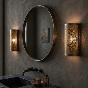 Op de afbeelding: Badkamer met een grote, ronde, goudomrande spiegel en twee bijpassende wandkandelaars. De wandkandelaars hebben een decoratief, geperforeerd ontwerp dat ingewikkelde lichtpatronen werpt. De wastafel heeft een zwart aanrechtblad en kraan.