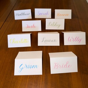 May include: White place cards with names written in various colors, including Matthew, Sarah, Bonnie, Josh, Libby, Charlotte, Lauren, and Willy. Two additional cards read "Groom" and "Bride". The cards are arranged on a wooden surface.