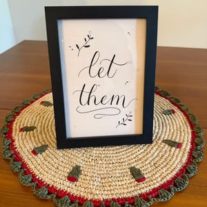 May include: A framed print with the words "let them" in elegant calligraphy. The black frame rests on a round, woven placemat with red and green trim and decorative accents. The placemat is made of natural fibers.
