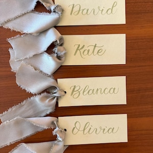May include: Four rectangular off-white paper tags, each featuring a name written in elegant gold cursive script. The names are David, Kate, Blanca, and Olivia. Each tag is attached to a light grey silk ribbon with frayed, raw edges.