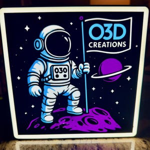 May include: A square, illuminated sign featuring a cartoon astronaut on a purple planet, holding a flag with "O3D CREATIONS" written on it. The astronaut is wearing a white and blue spacesuit. A purple planet with rings is in the background. The background is dark blue with white stars.
