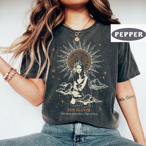 Pode incluir: Uma t-shirt cinzento-carvão de manga curta com uma impressão gráfica de uma pessoa ajoelhada sob um grande design de sol. A impressão inclui o texto THE SLAYER e um texto mais pequeno por baixo, rodeado por estrelas e nuvens.