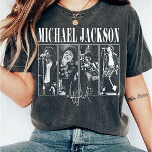 Puede incluir: Una camiseta de manga corta en color gris carbón con el texto MICHAEL JACKSON en letras mayúsculas blancas. Debajo del texto hay cuatro paneles fotográficos en blanco y negro de Michael Jackson actuando, con una firma impresa en la parte inferior.